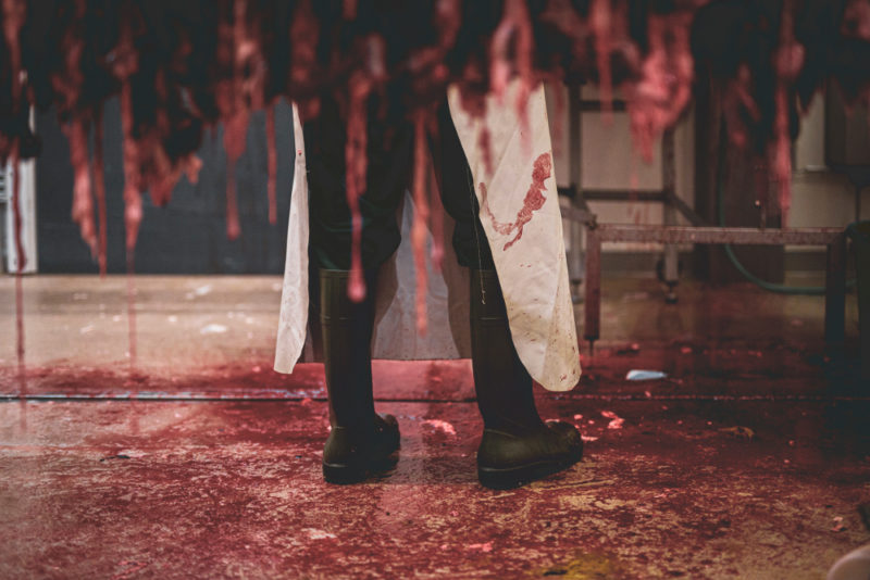 Teurastamon lattia on verinen ja siellä seisoo teurastaja mustissa kumisaappaissa.