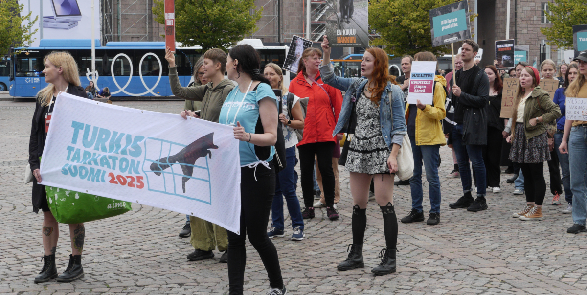 Joukko mielenosoittajia seisoo Rautatientorilla turkistarhauksen vastaisten kylttien kanssa.